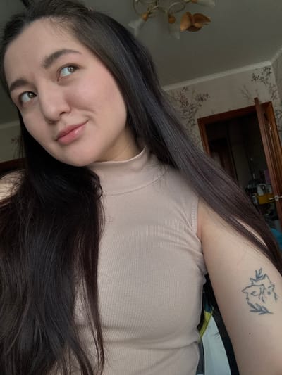 The image depicts a female individual, appearing to be a young adult, positioned in an indoor setting identified as a room within the city of Kurovskoye, Russia. She has long, dark hair, a light complexion, and light-colored eyes. She is wearing a sleeveless, light beige, ribbed mock-neck top. Her head is tilted slightly, and her gaze is directed upwards and to the viewer's right. A tattoo is visible on her left upper arm, depicting two stylized animal heads (resembling foxes or wolves) encircled by leafy branches. The background features a wall covered in patterned wallpaper with subtle floral or foliage designs. A dark wooden door, partially ajar, is visible on the right side of the frame, revealing a darker interior space beyond it. A multi-armed chandelier with light-colored elements is partially visible on the ceiling.