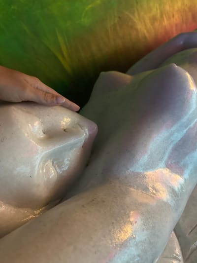 A close-up image shows a partially visible reclining white or light-colored statue of a human figure, possibly made of marble or similar material, being touched by a human hand with manicured nails. The head of the statue is prominent, showing facial features. The statue rests on a green and yellow surface. The location is Binh Lu, Vietnam.