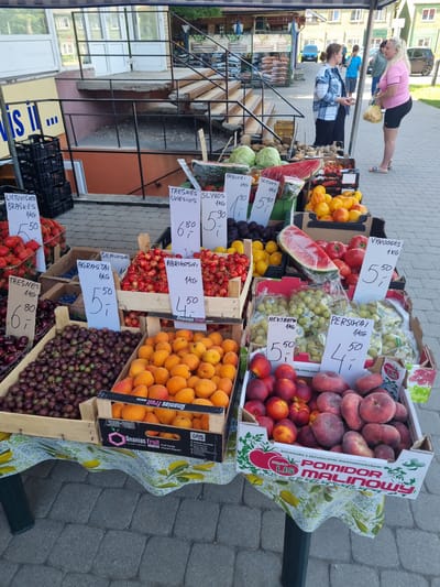 A fruit stand is set up outdoors in Kuršėnai, Lithuania. The stand displays a variety of fresh fruits, including strawberries, cherries, apricots, peaches, gooseberries, grapes, nectarines, and tomatoes. Some fruits are in crates, while others are packaged in plastic. Price tags are visible for some of the produce. In the background, there are people browsing the market and interacting with vendors. The setting appears to be a sunny day, with buildings and a cobblestone pavement visible.