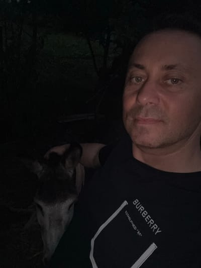 In the city of Tandzia, Georgia, a man is taking a selfie with a deer at night. The man is wearing a black t-shirt with a Burberry logo. The deer is looking at the camera, and the man is smiling. The background is dark, with trees and foliage visible. The image is taken in an outdoor setting.