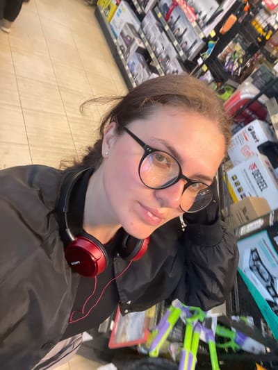 A young woman wearing round, black-rimmed glasses and red Sony headphones around her neck takes a selfie in a retail environment. She is dressed in a dark-colored jacket. Shelves filled with various products are visible in the background. The location is likely a retail store in Molokovo, Russia, based on other associated data. The floor has light-colored wood-look planks. There are water guns and boxes of DieReel on the shelf behind her.