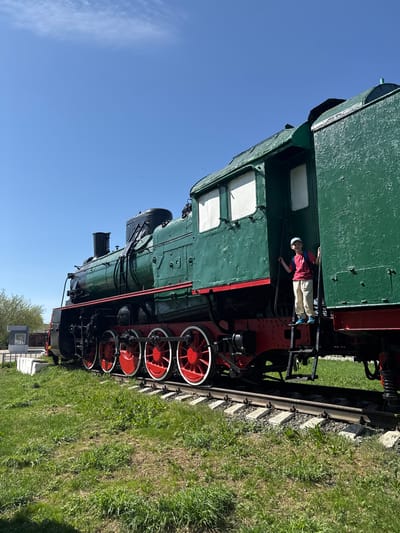 The image shows a green steam locomotive with red wheels, situated on a track in a grassy area under a clear blue sky. A young person wearing a red shirt, tan pants, and a light-colored cap is standing on the side of the locomotive. In the background, there is a small building and some trees. The location is likely Balezino, Russia, based on the prompt.