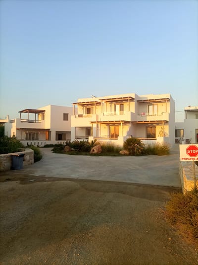 In the city of Naxos, Greece, the image captures a modern architectural complex bathed in the golden light of a sunset. The buildings, painted white with exposed wooden beams on their balconies, present a clean and minimalist aesthetic. They are multi-story structures with multiple balconies and large glass doors, suggesting they are likely apartments or a small hotel.
In the foreground, a paved driveway curves around to the buildings. To the left, a dark blue garbage bin sits beside a low stone wall. Further into the scene, a variety of drought-tolerant plants and grasses are arranged around the buildings, some with large rocks interspersed among them. A sign with a red stop symbol and the words "PRIVATE A" is prominently displayed on the right, indicating restricted access. The sky above is a clear, pale blue, transitioning to a warmer hue near the horizon. The overall impression is one of a serene and well-maintained property in a Mediterranean setting.