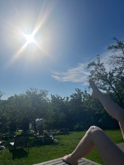The image captures a sunny day in Stăuceni, Moldova. The scene is set outdoors on a grassy area, likely a garden or park, with trees providing shade. In the foreground, parts of a person's legs and arm are visible, suggesting they are sitting or lying down on a wooden structure to the right of the frame. The individual's legs are bare and tanned, and a small white dot is visible on the knee, possibly a light reflection or a mark.
The sky is a clear blue, with a few wispy white clouds. The sun is a prominent feature, shining brightly with visible rays and lens flares, indicating it's likely midday or early afternoon. The lighting is strong, casting distinct shadows on the grass.
In the background, several lounge chairs are arranged on the lawn, some occupied by people. One person is partially visible sitting in a chair under a large umbrella. The overall atmosphere appears relaxed and leisurely, consistent with a warm, sunny day spent outdoors. The presence of lounge chairs and the overall setting suggest a recreational area, possibly a resort or a private garden. No text is visible in the image.