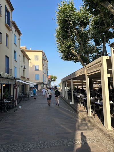The image depicts a lively street scene in Collioure, France, likely during the daytime. The architecture features pastel-colored buildings with blue shutters, characteristic of the town's Mediterranean style. On the left, a building houses a "Glacier" (ice cream shop) and other businesses with outdoor seating. To the right, a covered outdoor dining area of a restaurant is visible, with tables set with blue and white checkered cloths. Several people are walking along the cobblestone street. Some are casually dressed, suggesting a tourist area. The sun casts long shadows, indicating it might be late afternoon. The overall atmosphere is one of a bustling, charming European coastal town.
