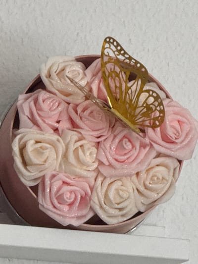 The image displays a close-up, elevated view of a decorative arrangement located in Vellmar, Germany. A round, light pink container, possibly a hatbox or gift box, is filled with approximately 12 artificial roses. The roses alternate in color between a soft, glittery light pink and a glittery off-white or cream. A golden, filigree butterfly decoration with openwork wings is affixed to the top of the roses, positioned towards the upper right of the container. The container rests on a clean, white horizontal surface, likely a shelf, which occupies the bottom portion of the frame. The background consists of a light-colored, possibly textured wall. No people or actions are depicted.