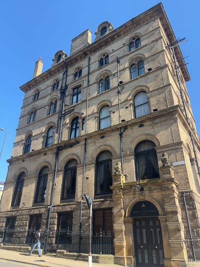 The image shows a large, historic building in Bradford, United Kingdom, located at 53.792 latitude and -1.751 longitude. The building is constructed of stone and has multiple stories, with a mix of arched and rectangular windows. Several decorative stone statues adorn the facade. The building's entrance is marked with a sign reading "Great Victoria Hotel". A single individual, a man wearing a blue jacket, walks on the sidewalk in front of the building. The scene is taken on a sunny day with a clear blue sky. There are no other people present. The ground is paved, with a curb separating the sidewalk from the street. The road is empty and appears to be a small street with a black street sign that reads "Granby St". A black metal fence separates the sidewalk from the building. The image depicts the exterior of a historical hotel, possibly a landmark in Bradford.