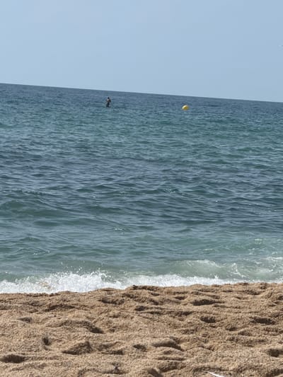 A wide-angle, eye-level shot captures a serene beach scene in Badalona, Spain. In the distance, a lone figure stands on a paddleboard in the calm, blue-green ocean under a clear, pale blue sky. Further out, a bright yellow buoy floats on the water's surface. The foreground is dominated by the textured, light brown sand of the beach, with gentle waves lapping at the shore, creating a line of white foam. The horizon where the sea meets the sky is a sharp, distinct line. The overall impression is one of tranquility and a quiet day at the beach.