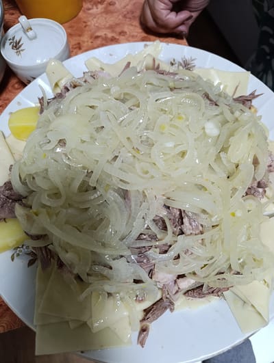 The image shows a large white plate filled with a dish consisting of wide, flat noodles, shredded meat (likely boiled), boiled potatoes, and a heaping pile of translucent, cooked onion slices. A small white ceramic bowl with a floral pattern is visible in the upper-left corner of the image. The background shows a table with a reddish-brown surface. A hand is visible in the upper-right corner.
The photograph was taken in Новоалександровка, Russia.
