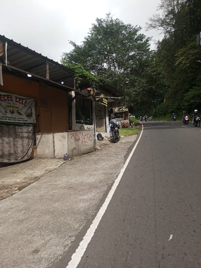 A roadside scene depicts a gray asphalt road curving right into the distance, with a solid white line defining its left edge. On the left, a concrete path runs alongside a row of rudimentary commercial structures with corrugated metal roofs. The leftmost structure features an orange wall and a hanging white banner with text "PECEL LELE" and "CAWANG" (Indonesian language), depicting chickens and fish, with a dark fabric draped below. The adjacent structure, partially concrete, exhibits extensive red and black graffiti. A black motorcycle is parked on the concrete path in front of this structure, with a black backpack on the ground beside it. A person in a dark shirt stands near the motorcycle. In the distance on the road, approximately six motorcycles with riders wearing helmets are traveling away from the viewer. The background on the right and center is dominated by dense, dark green trees, with a large, lighter green-leafed tree prominent behind the structures. The sky is overcast and uniformly gray.