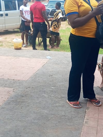 An outdoor scene in Ajalli, Nigeria, depicts five individuals and two vehicles on a paved and grassy surface under daylight conditions.
In the immediate foreground, a person is partially visible from the chest down, positioned on the right. This individual wears a yellow short-sleeved top with blue trim, black trousers, and orange flip-flops, displaying light blue nail polish on the toes. A black shoulder bag hangs on their left side. The ground beneath is a mix of textured grey and reddish-brown pavement.
In the mid-ground, four individuals are clustered near a patch of grass. From left to right:
1. A woman stands facing forward, smiling slightly. She wears a white t-shirt with "YES" and "USA" text, a dark pleated skirt, and white Adidas slide sandals. A brown shoulder bag is slung over her right shoulder. Next to her, on the ground, is a white lidded plastic bucket containing a yellowish substance.
2. A man stands with his back to the viewer, wearing a red t-shirt and black trousers. He is positioned to the left of a wooden wheelbarrow.
3. A person in a patterned gold and black short dress or tunic stands to the right of the wheelbarrow, facing the man in red. They have their right hand on their hip and wear pink flip-flops.
The wheelbarrow, with a single wheel, appears to contain unidentifiable items.
In the background, a white passenger van is parked on the far left. Further right and behind the mid-ground figures, a dark-colored SUV is partially visible. Green vegetation is present behind the wheelbarrow and the figures in the mid-ground. A crumpled white object lies on the paved ground in the foreground.