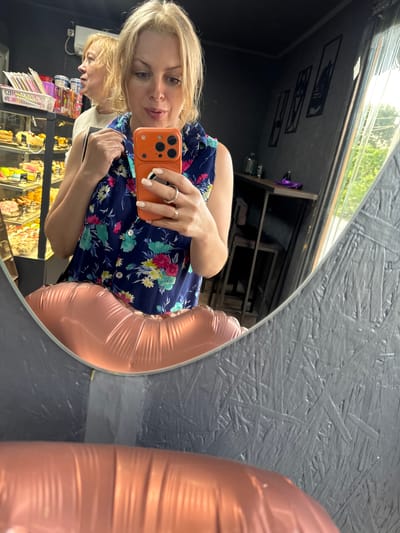 The image shows a woman taking a selfie in a bakery. She is wearing a floral print shirt and holding an orange phone. In the background, a display case filled with pastries is visible, as well as another woman. A large, rose-gold balloon is partially visible in the foreground, reflecting in a mirror. The setting appears to be a commercial establishment with dark, textured walls and a glass window showing greenery outside.