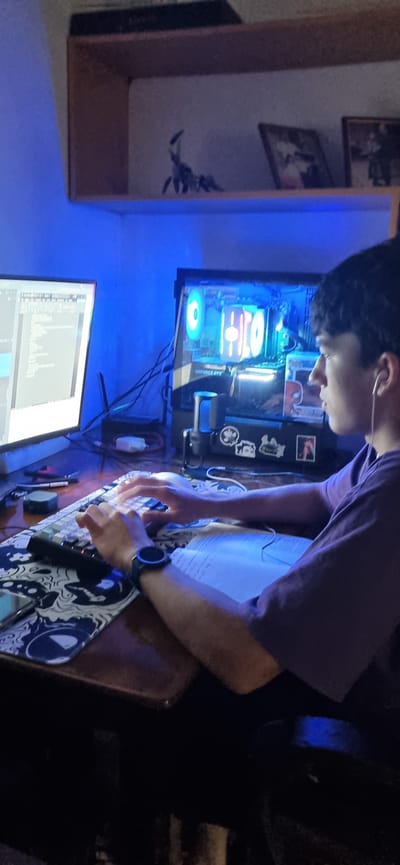A medium shot shows a young person with dark hair and earphones, wearing a purple t-shirt, sitting at a desk and typing on a keyboard. The person is looking at a computer monitor displaying code or text. To the right of the monitor is a glowing computer tower with internal lights. The desk is cluttered with a keyboard, a mousepad with a black and white abstract pattern, a watch on the person's left wrist, and an open book. Behind the desk, a shelf holds a decorative scorpion figurine and two framed pictures. The room is lit by blue ambient lighting, likely from the computer components and possibly LED strips. The scene appears to be a personal workspace, possibly a bedroom, in a private residence. The provided context indicating the location as Blizhnee, Ukraine, is a specific detail of the environment.