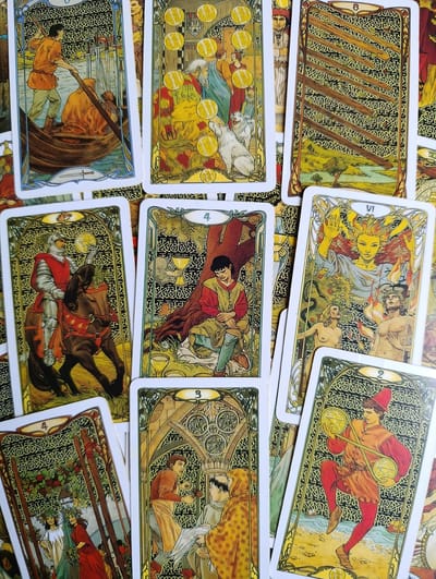 The image displays a collection of tarot cards laid out on a surface. The cards are intricately illustrated in a vintage style, featuring various figures and symbolic imagery. The depicted scenes include people in medieval or fantasy attire, horses, boats, coins, and nature elements. The overall color palette is warm, with prominent use of gold, green, and red. The cards are arranged in a slightly overlapping manner, suggesting they have been used for a reading. No discernible urban environment or specific location in Alexandrov, Russia, is visible.