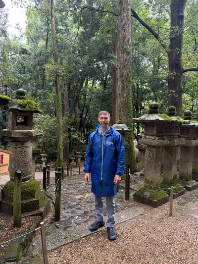 The image depicts a man standing on a path within the grounds of Kasuga Taisha Shrine in Nara, Japan, amidst numerous moss-covered stone lanterns and dense forest.
The central figure is an adult male with light brown, short hair, positioned facing the viewer. He is wearing a blue, knee-length rain jacket with a white logo on the left chest, light grey pants, and dark athletic shoes with teal accents. The ground beneath him is a mix of wet, uneven paving stones and light brown gravel, suggesting damp conditions or recent rain.
To the man's left, in the foreground, stands a large, tall stone lantern (tōrō) heavily covered in moss, particularly on its tiered roof and base. Vertical Japanese characters are inscribed on its main column, clearly showing "春日大社" (Kasuga Taisha) and "常夜燈" (Jōyatō - perpetual light). Small green plants grow from the top of this lantern. Partially visible behind it is a red and white wooden sign.
To the man's right, a cluster of three smaller, moss-covered stone lanterns stands on stone bases, each featuring a latticed section for light. Further into the background, additional stone lanterns are visible along a winding path. The entire scene is enveloped by a dense, lush forest featuring tall, straight tree trunks and a canopy of green foliage, indicating a humid, temperate environment. The light appears diffused, consistent with an overcast sky. Wooden and metal posts with thin wires line portions of the path, acting as a low barrier.