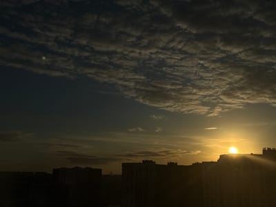The image captures a sunrise or sunset over an urban landscape in Kraskovo, Russia. The scene is dominated by a dramatic sky filled with textured clouds. In the upper half, dense, dark blue-gray clouds with visible patterns stretch across the sky. Towards the lower right, a brilliant golden sun is visible, partially obscured by the silhouette of a building. Strong rays of light emanate from the sun, illuminating the surrounding clouds with warm, golden hues.

Below the horizon, the dark silhouettes of numerous multi-story residential buildings are visible, stretching across the lower portion of the frame. These buildings are heavily back-lit by the low sun, rendering their architectural details largely indistinct.

Notable details include a small, bright green circular spot in the upper left quadrant, likely a lens flare or sensor artefact. The overall impression is one of early morning or late evening, characterized by the striking contrast between the dark foreground and the brightly lit sky, highlighting the natural beauty of the celestial event over a human-made environment. There are no people visible, nor any discernible text.