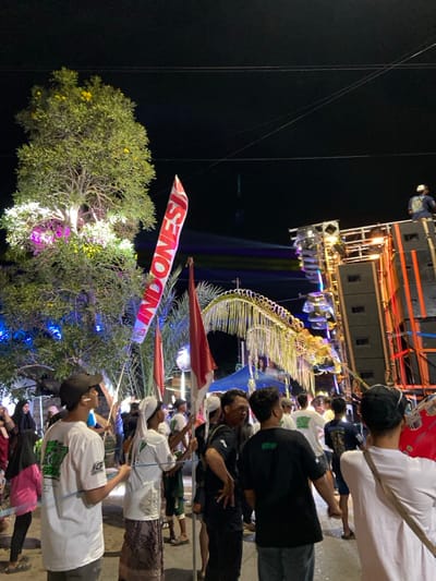 This image captures an outdoor nighttime event in Kedungbanteng, Indonesia. Numerous individuals, predominantly adult males, are gathered, facing generally towards the right side of the frame. Many wear casual attire, including t-shirts and caps, while some display white head coverings. In the immediate foreground, an individual with their back to the camera wears a white t-shirt and a dark baseball cap, with a light-colored strap across their shoulder.

A prominent vertical white banner, inscribed with "INDONESIA" in red capital letters, is held aloft by an individual in the left-midground, positioned next to a standard red and white Indonesian flag. Multiple similar red and white flags are visible throughout the crowd.

On the far right, a very large, multi-component sound system is erected, consisting of numerous black speaker cabinets stacked high, along with various stage lights and metal rigging. A lone individual is seated atop this tall structure. Beams of multicolored light emanate horizontally from the sound system, illuminating portions of the scene.

On the far left, a large tree with dense green foliage and some yellow blooms is illuminated from below by pink and white string lights. Above the crowd, a long, arcing decorative structure, appearing to be made of light-colored, possibly woven or ribbon-like materials, spans the mid-ground. The overall environment is dark due to the nighttime setting, with artificial lights providing illumination, and overhead utility lines are faintly visible against the dark sky. The ground appears to be a street or open area.