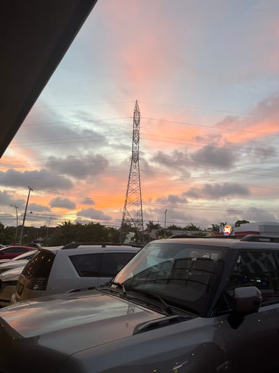 The image captures a dusky sky over Cancún, Mexico, with a prominent electrical tower in the mid-ground. The sky exhibits soft pastel colors, transitioning from pale blue at the top to shades of pink and orange near the horizon, indicative of sunset. Several power lines crisscross the frame, strung between poles visible in the distance and the large tower.

In the foreground, several vehicles are parked. The most visible is a dark gray SUV, its hood and windshield reflecting the sky. To its left, a portion of a white SUV is visible, and further to the left, the rear of a red car can be seen. Behind the vehicles, a treeline with palm trees suggests a tropical environment.

A Dairy Queen restaurant sign, with its distinctive red and blue logo, is perched atop a building in the background, to the right of the electrical tower. Streetlights and other utility poles are also present in the urban landscape. No people are clearly depicted in the scene. The overall impression is of a typical evening in a developed area within Cancún.