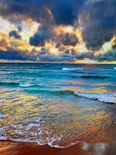The image depicts a serene coastal scene with a dramatic sky. Dark, heavy clouds are interspersed with vibrant shafts of golden light, suggesting either sunrise or sunset. The ocean is a deep, shimmering blue, with gentle waves rolling towards the shore. The water reflects the colors of the sky, creating streaks of gold and orange on the surface. The foreground shows a wet sandy beach, reflecting the light and the water. There are no people or man-made objects visible in the image. The scene is natural and tranquil. The location is identified as the city of Navahrudak, Belarus.