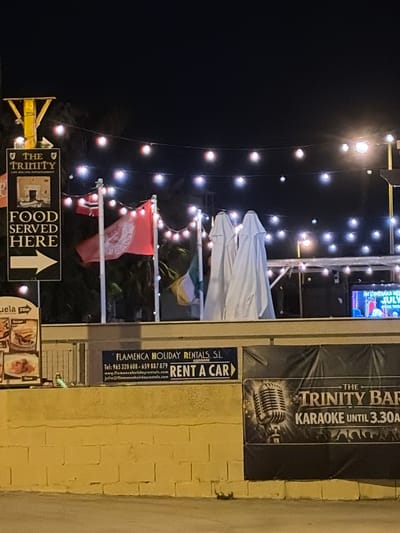The image depicts a nighttime scene outside a bar or establishment in Orihuela, Spain. In the foreground, a yellow brick wall is visible. Above the wall, a dark banner with a vintage microphone and text reading "THE TRINITY BAR KARAOKE UNTIL 3.30A" is displayed. To the left of this banner, a dark blue sign with white text advertises "FLAMENCA HOLIDAY RENTALS, S.L." and provides contact information. Further to the left, a tall, dark sign with white text reads "THE TRINITY" at the top, followed by "FOOD SERVED HERE" and a white arrow pointing to the right. The background is illuminated by numerous string lights, creating a festive atmosphere. Several flags are visible, including what appears to be a red and white Norwegian flag and a red flag with a white emblem, possibly a company logo. Two white, shrouded objects, likely umbrellas, stand next to the flags. In the far right of the image, a screen displays what looks like a colorful advertisement or event listing. The overall impression is of a lively entertainment venue at night.