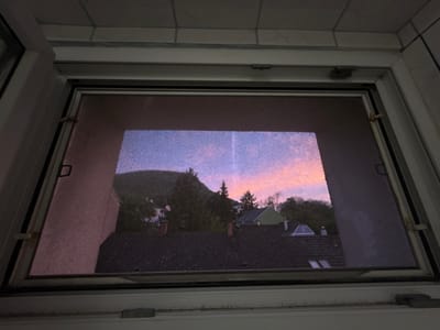 A view from inside a building, through a window with a screen, shows the town of Hainburg an der Donau, Austria, at dusk. The foreground is a window frame with a screen partially obscuring the view. Beyond the screen, rooftops of houses with varied shapes and colors are visible. Further in the background is a hillside with trees. The sky is a gradient of colors, transitioning from blue to pink as the sun sets. The image is taken at an oblique angle, looking slightly up and out. The lighting is dim, suggestive of late evening.