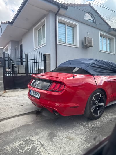 A red Ford Mustang convertible, partially covered with a dark blue car cover, is parked on a paved driveway in front of a grey, two-story house. The house has white-trimmed windows and a dark brown tiled roof. An air conditioning unit is mounted on the exterior wall. A black metal gate with vertical bars and a concrete post stands to the left of the car, with a mailbox attached to the post. The car's license plate reads "A 8008 PB". The ground is a rough, grey concrete surface, with some cracks and a slight incline. The sky is partly cloudy. The image appears to be taken in the city of Aytos, Bulgaria, based on the license plate format.
