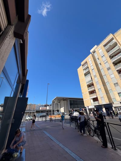 The image captures an urban street scene in Bari, Italy, under a bright, clear blue sky with a single small white cloud visible. The perspective is low, looking up at surrounding buildings and across a sidewalk.
On the left, a tall, modern apartment building dominates the view with its distinctive facade of alternating light-colored and beige horizontal stripes, featuring numerous windows and recessed balconies.
On the right, a busy sidewalk runs alongside a street. In the immediate foreground, part of a building with large, reflective windows is visible, topped by a dark brown architectural structure where the number "54" is faintly inscribed. Above these windows, a partial blue and yellow sign, possibly for a commercial establishment, can be seen.
Several people are present on the sidewalk. One person, dressed in dark attire, stands next to a bicycle, seemingly engaged with a phone or an item in their hand. Further down, another individual in a striped shirt is crouching near the reflective windows, appearing to organize or handle items. A third person, wearing light blue pants and a white top, walks along the paved sidewalk. In the background, other indistinct figures, street lights, and various traffic signs (including a triangular warning sign and circular regulatory signs) are visible, along with parked or moving vehicles and more urban buildings.
The bright sunlight suggests it is daytime, likely midday or early afternoon, with shadows cast on the pavement. The overall scene depicts a vibrant, contemporary urban environment.