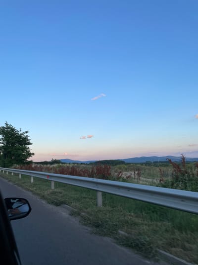 The image captures a view from a vehicle moving along a road, likely on the outskirts of Ovoshtnik, Bulgaria. A silver metal guardrail runs along the right side of the road, separating it from a grassy verge and a field of dry, reddish-brown plants. In the distance, rolling hills form a dark, silhouetted horizon under a clear blue sky with a few wispy clouds. The sky transitions to softer pinks and blues near the horizon, suggesting it is late afternoon or early evening. A large, leafy green tree stands to the left, closer to the road. Reflected in the car's side mirror, a person's hand, holding a phone with a white case, is visible. The road surface appears dark and asphalt.