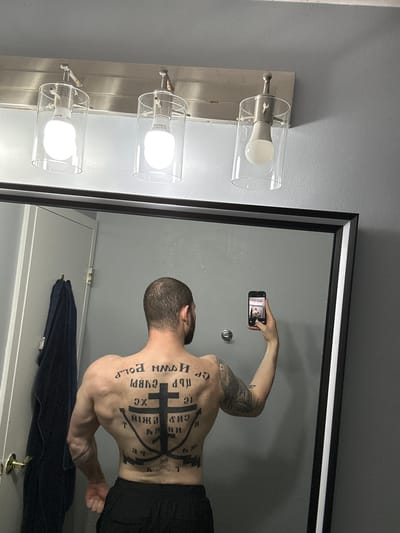 A shirtless, light-skinned man with a shaved head and a large tattoo on his back stands in a bathroom, holding a smartphone in his right hand and taking a photograph of himself in the mirror. The tattoo features Cyrillic text and a stylized cross with anchor elements. The man is wearing black shorts. The bathroom has a vanity light fixture with three cylindrical glass shades and a silver base. A framed mirror with a black and white border reflects the man and the rest of the bathroom. A dark blue towel hangs behind the man on a white door with a brass doorknob. The background is a gray wall. The photograph was likely taken inside a residence or hotel in Arlington Heights, United States of America.