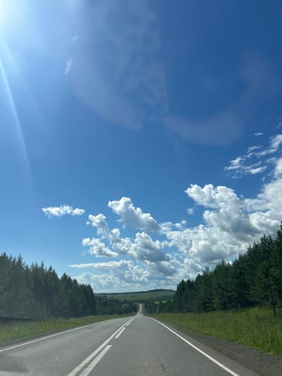 The image depicts a straight asphalt road stretching into the distance, flanked by dense coniferous trees on both sides. The road has white lane markings. The sky is a vibrant blue, dotted with fluffy white clouds. The sun is shining brightly, causing a lens flare in the upper left corner of the image. The overall environment suggests a rural or natural setting, possibly in or around the city of Golyanovo, Russia, given the context. There are no people or animals visible in the scene. The perspective is from the driver's or passenger's seat of a vehicle.