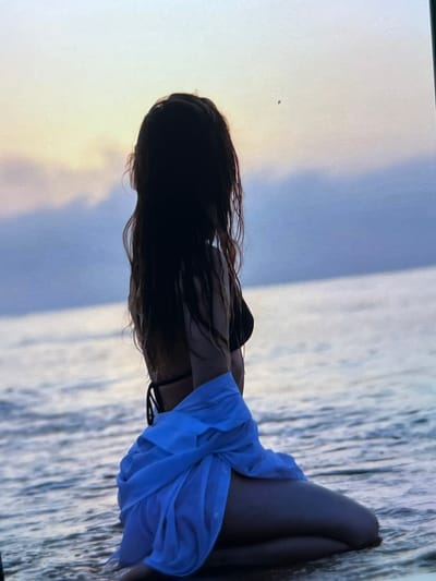 A person with long, dark, wet hair is kneeling in shallow water at the edge of a body of water, possibly the sea or a large lake. They are facing away from the camera, looking towards the horizon where the sky transitions from a pale yellow near the horizon to a muted blue above. The person is wearing a black bikini top and a light blue garment wrapped around their lower body. The water is calm with gentle ripples. The overall scene suggests a serene, possibly sunset or sunrise atmosphere. The location is not specified in the image and cannot be determined from visual cues.