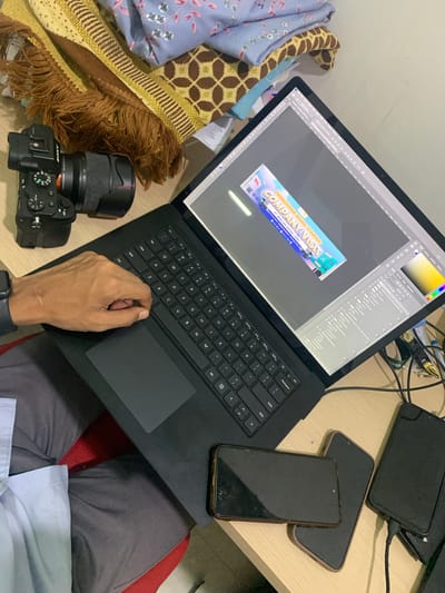 An overhead shot shows a laptop with a graphic design program open, displaying a banner for "COMPANY VISIT". A Sony camera with a lens is to the left of the laptop. A person's hand rests on the laptop keyboard. Three smartphones are on the desk, two are stacked and one is charging. The scene is indoors, with a patterned cloth in the background. The overall scene suggests a creative workspace in Parung Panjang, Indonesia.