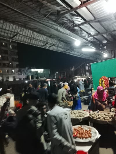 Under a high, industrial-style roof with bright lighting, a bustling outdoor market scene unfolds at night. Numerous people mill about, examining goods displayed in large, round baskets. Several vendors and customers are visible, some wearing traditional clothing. Buildings in the background are blurred, suggesting a densely populated area. The market appears active and crowded, with an assortment of produce and other items for sale.