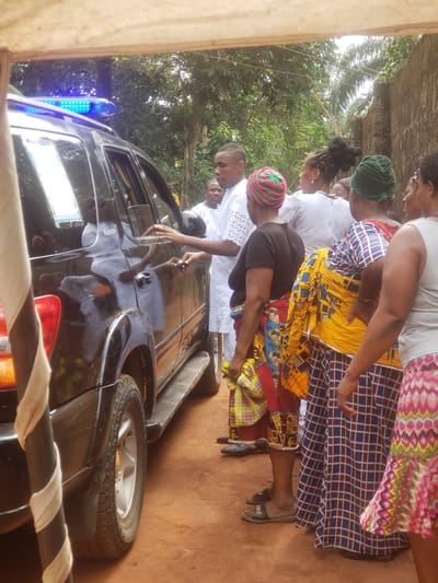 This photograph depicts a gathering of people, primarily women and two men, around a black car in an outdoor setting in Umundugba, Nigeria. The scene unfolds on reddish-brown earth, with lush green trees and foliage in the background, along with a rustic wall.
In the foreground, several women are visible in colorful attire. One wears a grey top and a vibrant, patterned skirt with pink, yellow, and green hues. Another has a green headwrap and a plaid blue and white skirt, accessorized with a yellow patterned fabric around her waist. A third woman features an elaborate dark hairstyle and a white eyelet lace top. A fourth woman wears a distinctive red, green, and white striped cap and a black top.
A man in a white lace top or agbada stands beside the black car, reaching towards its door, possibly opening or closing it. Another man is partially visible behind him. The car, which appears to be an SUV or a similar utility vehicle, has a blue flashing light on its roof, suggesting it might be an emergency, official, or ceremonial vehicle. A spare tire is mounted on the rear of the car.
The activity appears to be a social or community event, possibly centered around the car or an arrival/departure. The formal and colorful traditional clothing worn by the individuals suggests a special occasion. The time of day is daylight, with bright, natural illumination. No specific text is clearly legible on the car's plate or elsewhere, apart from a partial word "IOFU" on a slipper. The overall mood seems attentive and communal, characteristic of a significant local event.