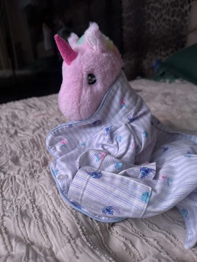 A close-up, low-angle shot shows a pink plush unicorn toy sitting on a white textured surface, possibly a bed. The unicorn is wearing a light blue and white striped pajama shirt with a pastel floral and Stitch pattern. The unicorn's head is turned to the left, and its horn is pink. The background is dark and blurred, with a hint of what appears to be a dark, patterned fabric on the right. The scene does not provide any information or context to place it in Slanchevo, Bulgaria.