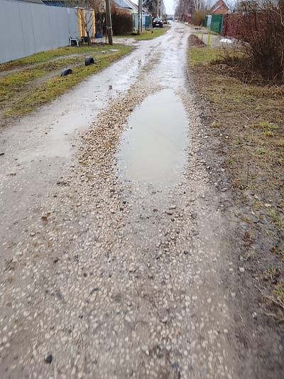 The image depicts a waterlogged dirt road in what appears to be a residential area in Высокиничи, Russia. The road surface is primarily gravel and dirt, with several puddles of varying sizes. Two black tires are visible on the grassy verge to the left of the road. In the background, houses and fences are visible, along with utility poles. The scene suggests a rural or semi-rural environment, possibly after recent rainfall.