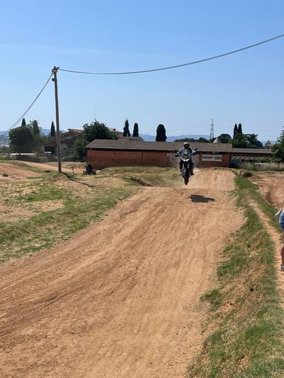 The image shows a dirt road in l'Ametlla del Vallès, Spain. A motorcyclist is riding down the road, kicking up dust. Buildings are located on the left and green grass on the right. A utility pole with wires is on the left side of the road. The sky is clear and blue.