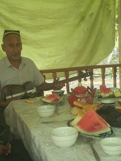 An elderly man wearing a traditional Uzbek skullcap sits at a table covered with a white lace tablecloth. He is holding and playing a stringed instrument, possibly a dutar, and is looking towards the viewer. On the table in front of him, there are several white bowls, slices of watermelon, bread, and a red teapot decorated with floral patterns. The scene appears to be set outdoors, possibly on a porch or under a canopy, with a blurred natural background suggesting greenery and possibly a body of water. The man's attire and the presence of the instrument suggest a cultural or musical context. The setting of Avval', Uzbekistan, suggests a cultural event or a gathering in a rural or semi-rural area.
