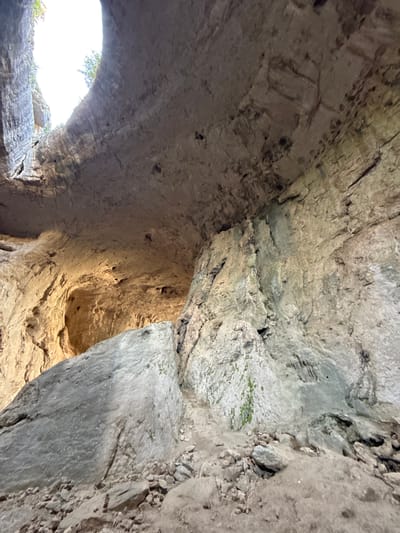 The image depicts the interior of a large cave, likely a natural rock formation. The cave ceiling is a prominent feature, appearing smooth and arched, with light filtering in from an opening at the top. The walls are rough and textured, displaying layers of rock and numerous small holes or indentations. Large boulders and rock debris are scattered across the cave floor, suggesting a natural and undeveloped environment. The lighting suggests it is daytime, with sunlight illuminating parts of the cave's interior, creating areas of shadow and highlighting the rock formations. The scene is devoid of any human presence or activity. This imagery is consistent with the geological features found in the karst region of Karlukovo, Bulgaria, known for its extensive cave systems and natural arches, such as the "Eyes of God" rock formation.