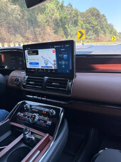 The image provides a driver's-side perspective from inside a modern car, looking out the windshield and across the dashboard. The car's interior features a luxurious design with brown leather-textured material on the dashboard and door panels, complemented by dark wood trim and chrome accents.

A large, vertically mounted infotainment screen dominates the center of the dashboard. It displays a navigation map, identifying the route as "MEX-95D / México - Cuernavaca" and "MEX-95 / Paso Expréss", aligning with the Coajomulco, Mexico location. The screen shows a current time of "2:15" and an outside temperature of "28°". A notable alert states "Vehículo detenido En 410 m" (Vehicle stopped in 410m), with options "Sigue ahi" (Continue there) and "No está ahi" (Not there), suggesting a road incident or traffic stop ahead. Other visible text on the screen includes "Phone List", "Climate", "Vehicle Settings", "Apps", "Settings", "Features", "Audio", and "Apple CarPlay". A speed limit of "90" is shown on the map, and a distance of "20 km" is also displayed. Below the screen, the gear selector indicates 'D' for Drive.

To the right of the screen, the center console houses climate controls with various buttons and knobs, labeled "TUNE", "REAR", "ON", "PASSENGER AIRBAG", "REAR A/C", "MAX A/C", "MENU", "AUTO", and "VOL". A USB charger or similar device is plugged into a compartment within the console.

Beyond the windshield, a bright, sunny day with a clear sky is visible. The car is on a highway, with lush green trees and foliage covering a hillside. A yellow guardrail runs alongside the road. Two yellow chevron signs, indicating a left curve, are prominently displayed, one in the mid-ground and another further down the road. The scene suggests the car is either in motion or temporarily stopped on a scenic, possibly mountainous, highway.