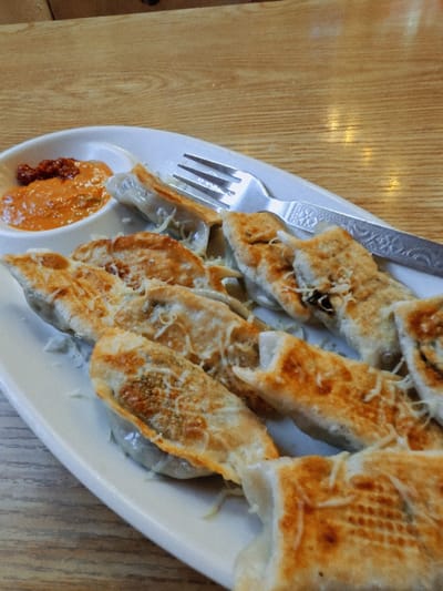 A plate of pan-fried dumplings, topped with shredded cheese, sits on a wooden table, accompanied by a small dish of dipping sauce. A silver fork rests on the plate. The scene appears to be set in a restaurant within Kathmandu Metropolitan City, Nepal.