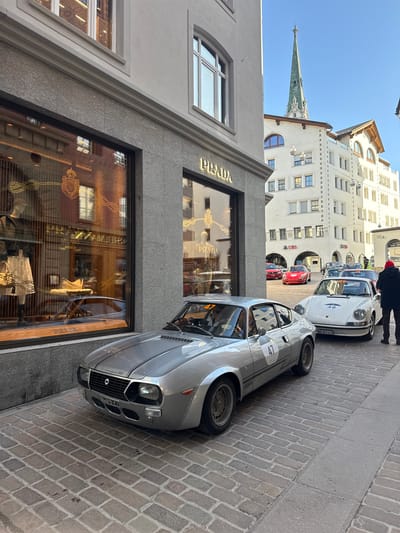 The image depicts a street scene in Sankt Moritz, Switzerland, characterized by classic cars parked on a cobblestone road in front of luxury retail and traditional Alpine architecture.

In the foreground, a silver Lancia Fulvia Sport Zagato with racing number "47" on its side and front is parked diagonally on a cobblestone street. It features a black racing stripe over the hood and an Italian license plate, "S44975 E1". Adjacent and slightly behind it on the right, a white Porsche 911, bearing racing number "48" on its hood, is parked parallel to the Lancia. Its license plate reads "VD-321417". To the far right, a person in a dark jacket and a red beanie stands observing the vehicles, with their back mostly to the camera.

The left side of the scene is dominated by a luxury boutique, identified by the gold "PRADA" logo above its display windows. The building facade at street level is clad in light grey granite, transitioning to light grey stucco or paint on the upper floors. The large display windows reflect the opposite buildings and showcase luxury items and mannequins.

In the mid-ground, a line of other vintage and classic cars is visible, including a red Lancia Stratos and a red Porsche, parked further down the street, suggesting a rally or automotive event. A multi-story, light-colored building with traditional arched windows and exposed timber eaves, prominently featuring a "UBS" logo at ground level, forms the backdrop for these vehicles. Decorative string lights with lantern-like fixtures hang across the street in this area. Another individual, possibly event staff, is visible standing near the parked cars in the mid-ground.

Above the buildings, a tall, slender church spire with a green-patinaed metal roof and gothic architectural details rises sharply against a clear, bright blue sky. The overall lighting suggests strong sunlight. The street surface throughout the visible area consists of varied shades of grey and tan cobblestones, with a narrow, smoother concrete path visible on the immediate right.