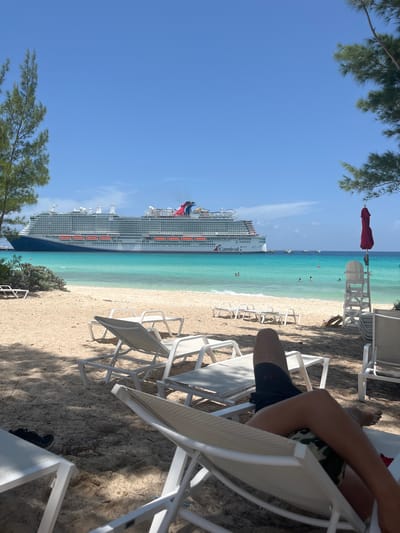 In the City of Freeport, Bahamas, a beach scene unfolds under a clear blue sky. The foreground features white lounge chairs on a sandy beach, with one chair occupied by a person from the knees down, wearing black shorts and a camouflage-patterned fabric. In the midground, a line of empty white lounge chairs stretches along the shore, leading to the ocean. The turquoise water is calm, with a few people swimming or wading. A large Carnival cruise ship is anchored offshore. To the right, a white lifeguard stand with a red umbrella is visible. The background is dominated by the vast blue sky and a few scattered clouds. The overall impression is one of a relaxing tropical vacation.