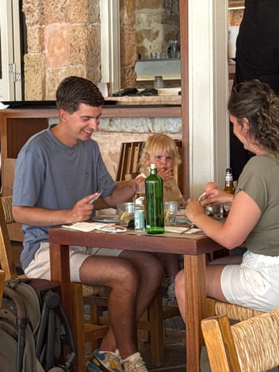 A family is dining at a table in a restaurant in Chania, Greece. A man wearing a blue t-shirt and white shorts is seated at the left of the table, holding a knife and fork, and smiling. A young child with blond hair is seated in front of him, eating. A woman with curly brown hair, wearing a green t-shirt and white shorts, is seated on the right side of the table, also eating. The table is set with plates, glasses, and a green bottle. A stroller is parked next to the table. The restaurant has stone walls and wooden furniture.