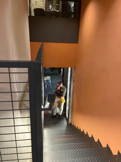 A man in a brown shirt and a yellow utility belt is standing at the top of a dark staircase. The man is looking down, his chin resting on his hand. His left leg is slightly bent, his right leg straight. The staircase leads down into a dimly lit space with a tiled floor. The walls are painted in various shades of orange. There is a black railing on the left side of the stairs. The image was captured in a building located in Las Palmas de Gran Canaria, Spain, at coordinates 28.102 latitude, -15.415 longitude. The scene is likely from a restaurant or a bar. The light comes from the top of the stairs.