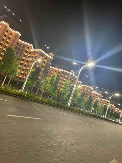 The image depicts a street scene at night in Grozny, Russia. The primary subjects are the illuminated buildings lining one side of a wide asphalt road. The buildings are modern, with a warm, orange glow emanating from their exterior lights, highlighting their architectural details, including arched windows and decorative cornices. Trees are planted along the sidewalk in front of the buildings, their green foliage providing a contrast to the warm building lights. Streetlights with bright, white lights are positioned along the road, casting beams that create a dramatic effect against the dark sky. The road itself is paved and marked with white lines, suggesting it is a well-maintained urban thoroughfare. The perspective suggests the photo was taken from a moving vehicle, with the road streaking past. The time of day is clearly night, and the weather appears clear, with no indication of rain or fog. The overall impression is one of a vibrant and well-lit urban environment at night. There is no visible text in the image.