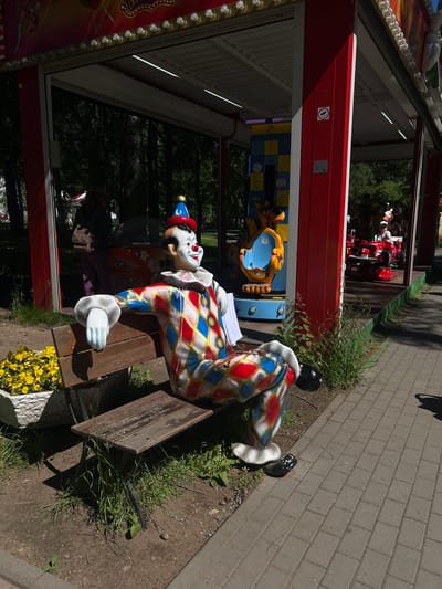 The image depicts an outdoor amusement park scene in Zelenogorsk, Russia. In the foreground, a life-sized clown statue is seated on a wooden bench. The clown wears a colorful diamond-patterned costume in red, blue, and white, a red and blue cone hat, and has a painted white face with a red nose and smile. The bench is weathered and surrounded by sparse green grass and a planter filled with yellow flowers. Behind the clown, a red and white amusement ride structure is visible, with children on the ride in the background. The ground is a mixture of dirt and a paved walkway made of grey rectangular bricks. The scene is illuminated by sunlight, casting shadows.
