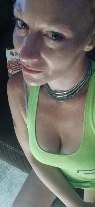 The image depicts an adult female in a close-up, high-angle portrait, likely a self-portrait, in an indoor setting.
**People:** One female individual is visible. She has light skin, green eyes, and appears to be looking upwards and slightly to the left. Reddish discoloration is present around her left eye and on her forehead. She is wearing a neon green tank top and a silver-toned, multi-strand choker necklace. A dark mark or small tattoo is visible on her left upper arm.
**Objects:** Behind and to the left of the subject's head, a partially visible box of "H-E-B Toasted Pastries Frosted Strawberry" is present, showing the number "12" (likely indicating quantity). A dark, upholstered surface, possibly a couch or chair, with visible stitching, is in the left foreground. A light-colored, possibly tiled or concrete floor is visible in the lower left.
**Actions:** The subject is posing for the camera.
**Environment:** The scene is indoors, with lighting that appears somewhat dim, featuring a light source positioned to illuminate the subject from above and front.
**Location:** There are no specific visual identifiers within the image (e.g., landmarks, street signs, unique architecture) to confirm its location in Santa Fe, United States. The H-E-B brand is a regional grocery chain, but its presence does not specifically pinpoint Santa Fe.