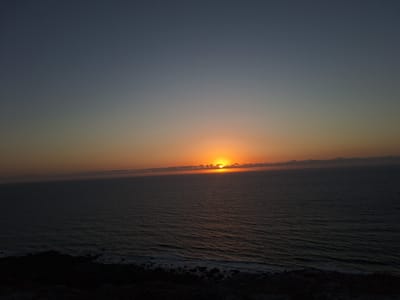 The image depicts a sunset over the Atlantic Ocean, viewed from a rocky cliffside. The sky is a gradient of dark blue at the top, transitioning to hues of orange and yellow near the horizon where the sun is setting. The sun itself is partially obscured by thin clouds. The ocean is dark and appears to have moderate waves crashing against the shore. The foreground shows a dark, rugged cliff with scattered rocks. The overall scene is one of natural beauty and tranquility. The location is identified as Ahrara, Morocco. There are no people or identifiable structures visible in the image.