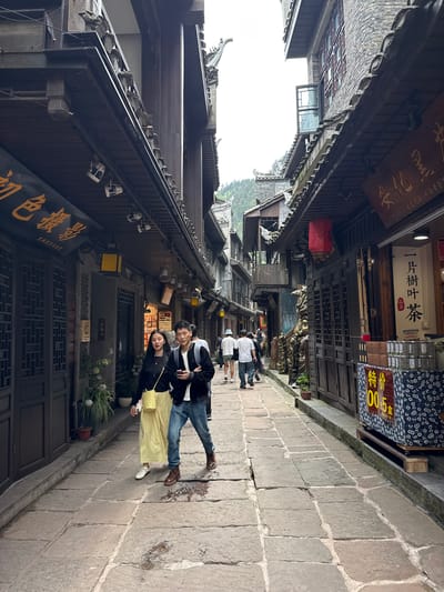 Here's a detailed description of the image:

**Scene:** The photo shows a narrow street in the city of Fenghuang, China. The street is paved with large, irregular stone slabs. Traditional Chinese buildings line both sides of the street. These buildings are predominantly made of dark wood, with intricate architectural details, overhanging eaves, and traditional tiled roofs.

**People:** In the foreground, a couple is standing and smiling at the camera. The woman is wearing a black jacket and a long yellow skirt. The man is wearing a black and white jacket and jeans. Further down the street, other people are walking away from the camera.

**Objects and Details:**

*   Signage: Various signs with Chinese characters are visible on the buildings. One sign reads "初色援影" (Chu Se Yuan Ying), indicating a photography studio offering traditional ethnic clothing rental and photo services.
*   Shops: One shop sign reads "安化黑 古文毛尖海西黄金" (Anhua Hei Guwen Maojian Haixi Huangjin) and "一片樹叶茶 品茶" (Yī piàn shù yè chá pǐn chá), referring to tea products being sold.
*   Lanterns: A red lantern hangs from one of the buildings.
*   Merchandise: A sign displaying "特价 00:5盒" (Tejia 00:5 He) is visible, suggesting a special price on a boxed item, potentially tea, at ¥55.00
*   Plants: Plants are seen in pots outside the building.

**Environment:** The sky appears overcast or slightly foggy, creating soft, diffused lighting. The architecture suggests a historical or tourist-oriented area. The overall atmosphere is one of a charming, traditional Chinese street scene.